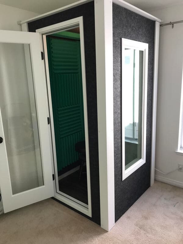 Soundproof recording booth with glass door, green acoustic panels, and carpeted floor.