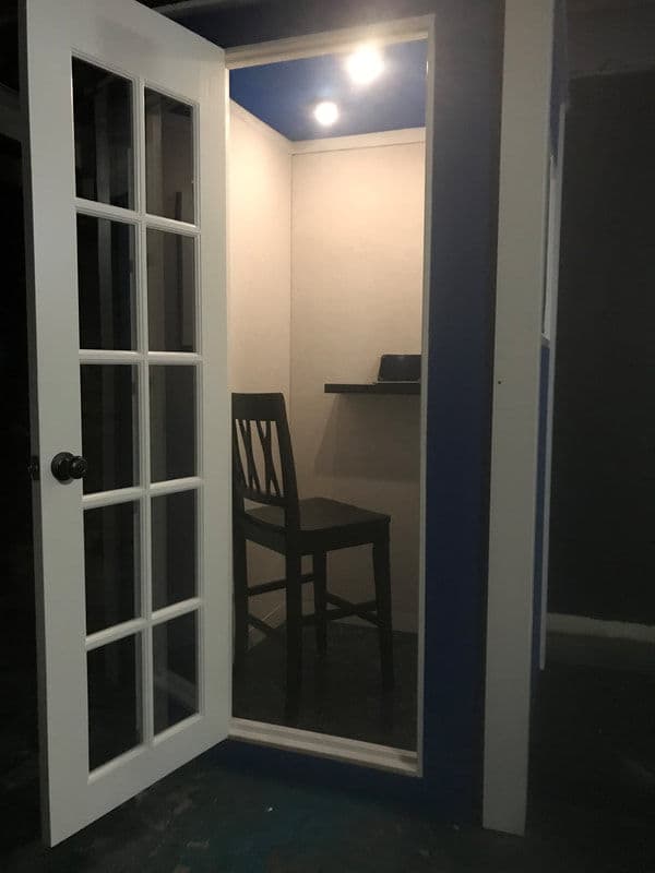 Interior of a modern sound booth with a chair and shelf, featuring glass-paneled door.