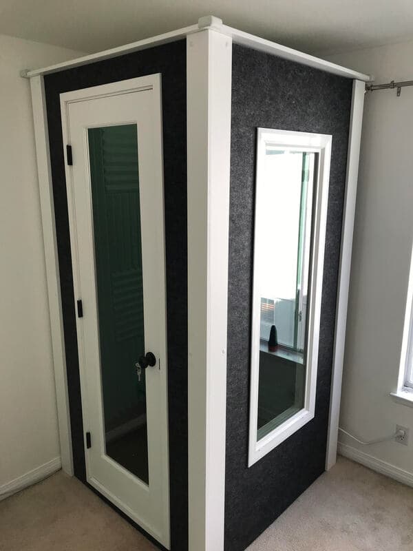 Soundproof recording booth with glass window and white door in a room setting.
