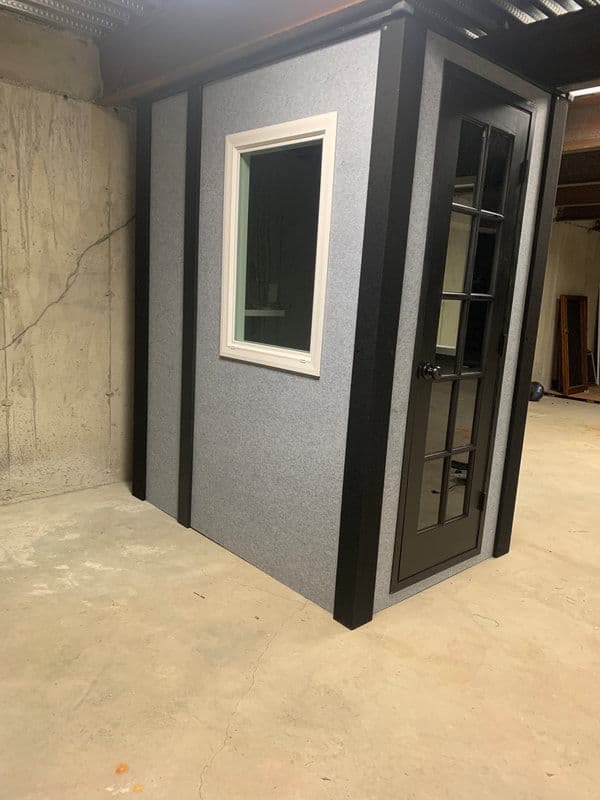 Modern soundproof booth with glass door and window in a basement setting.