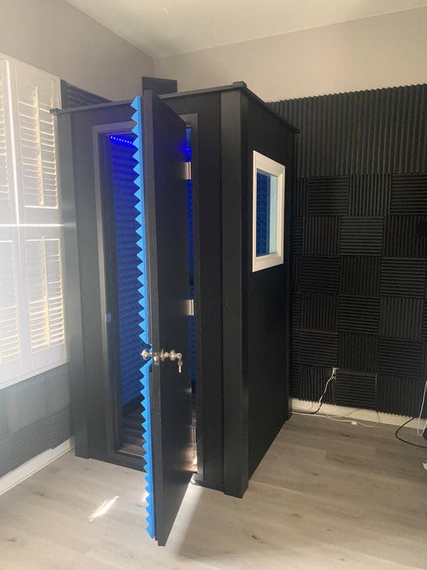 Soundproof vocal booth with blue lighting, situated in a room with acoustic panels.