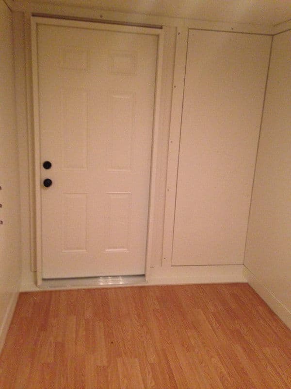 Interior view of a room with a white door and blank walls, featuring wood flooring.