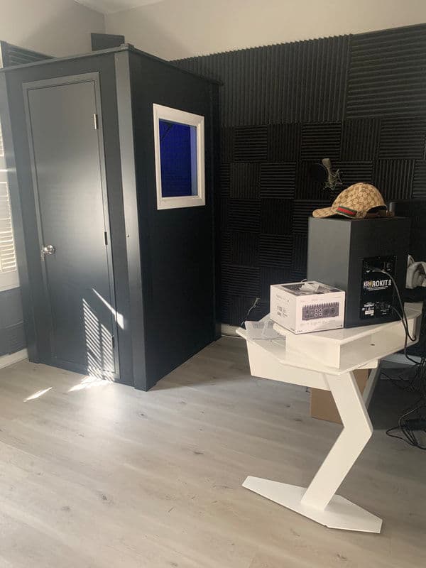 Modern recording studio with black soundproofing, a vocal booth, and white desk furniture.