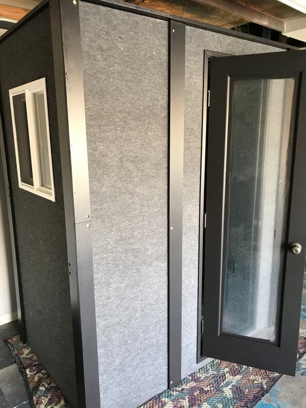 Soundproof booth with gray fabric exterior and glass door, designed for recording or privacy.