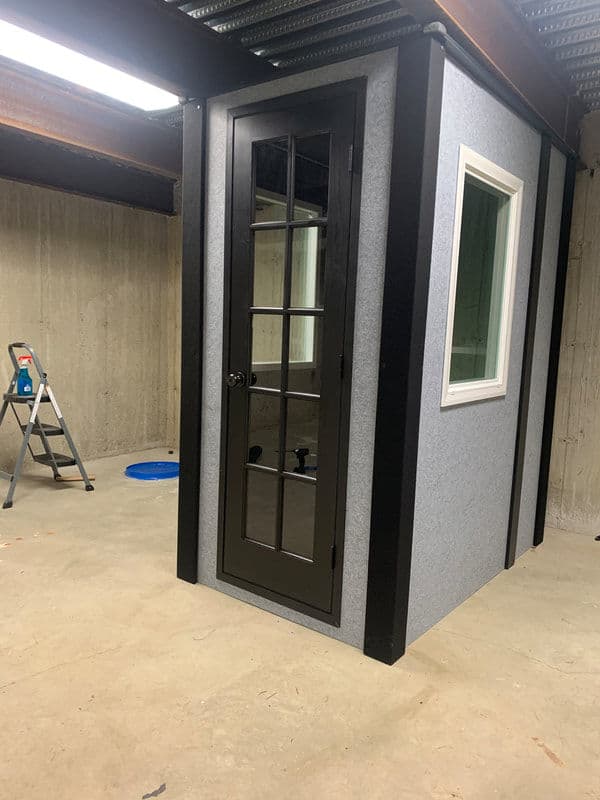 Interior view of a modern soundproof booth with a glass door and gray walls in a basement setting.