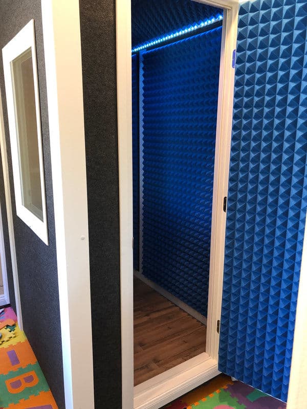 Soundproof recording booth with blue acoustic panels and a wooden floor, featuring a clear door.