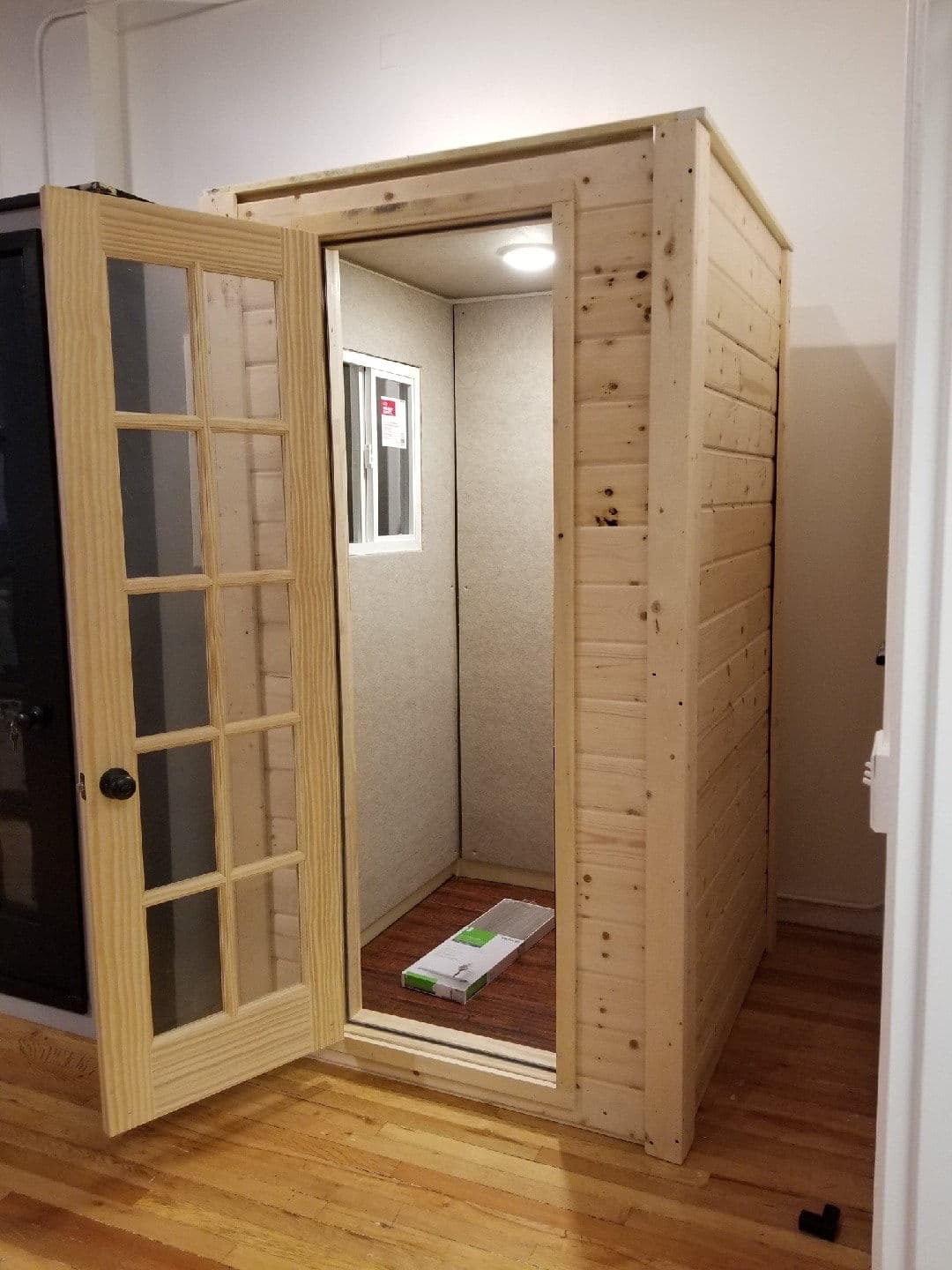 Wooden soundproof booth with glass door, open to reveal interior space and flooring.