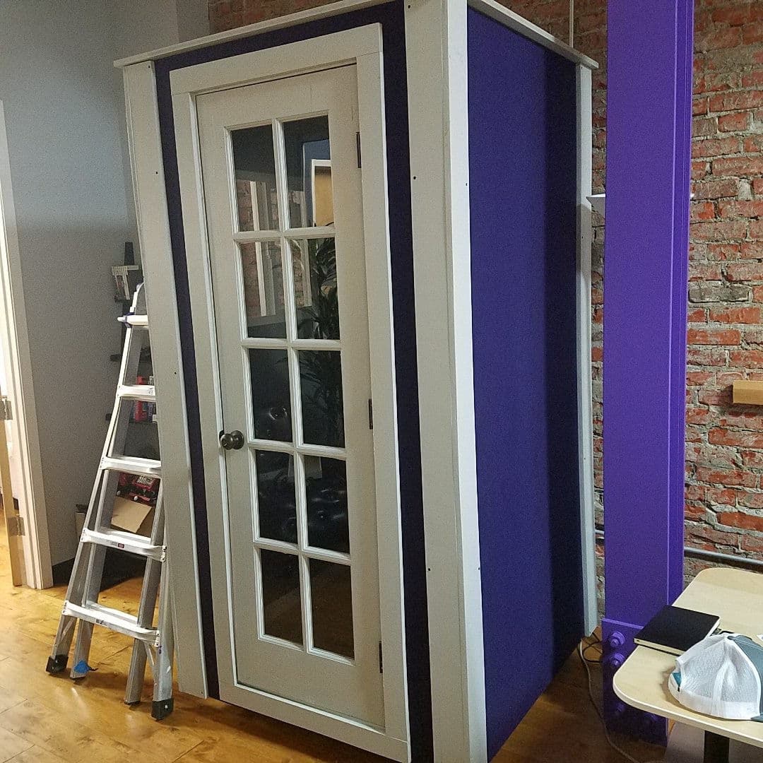 Soundproof booth with purple paneling and glass door, situated in a modern office space.